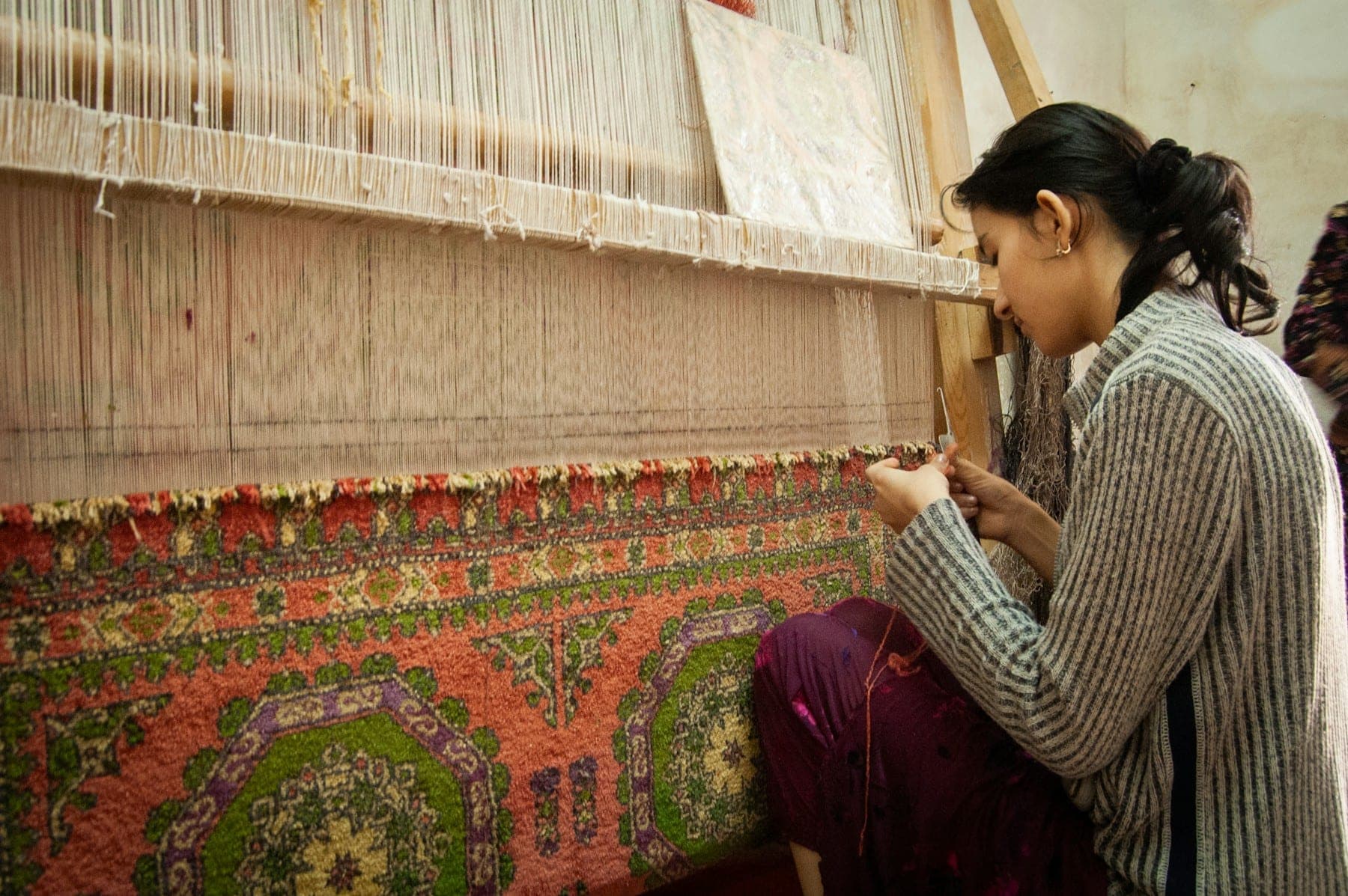 Weaver knotting pile on an upright loom beneath rows of warp threads