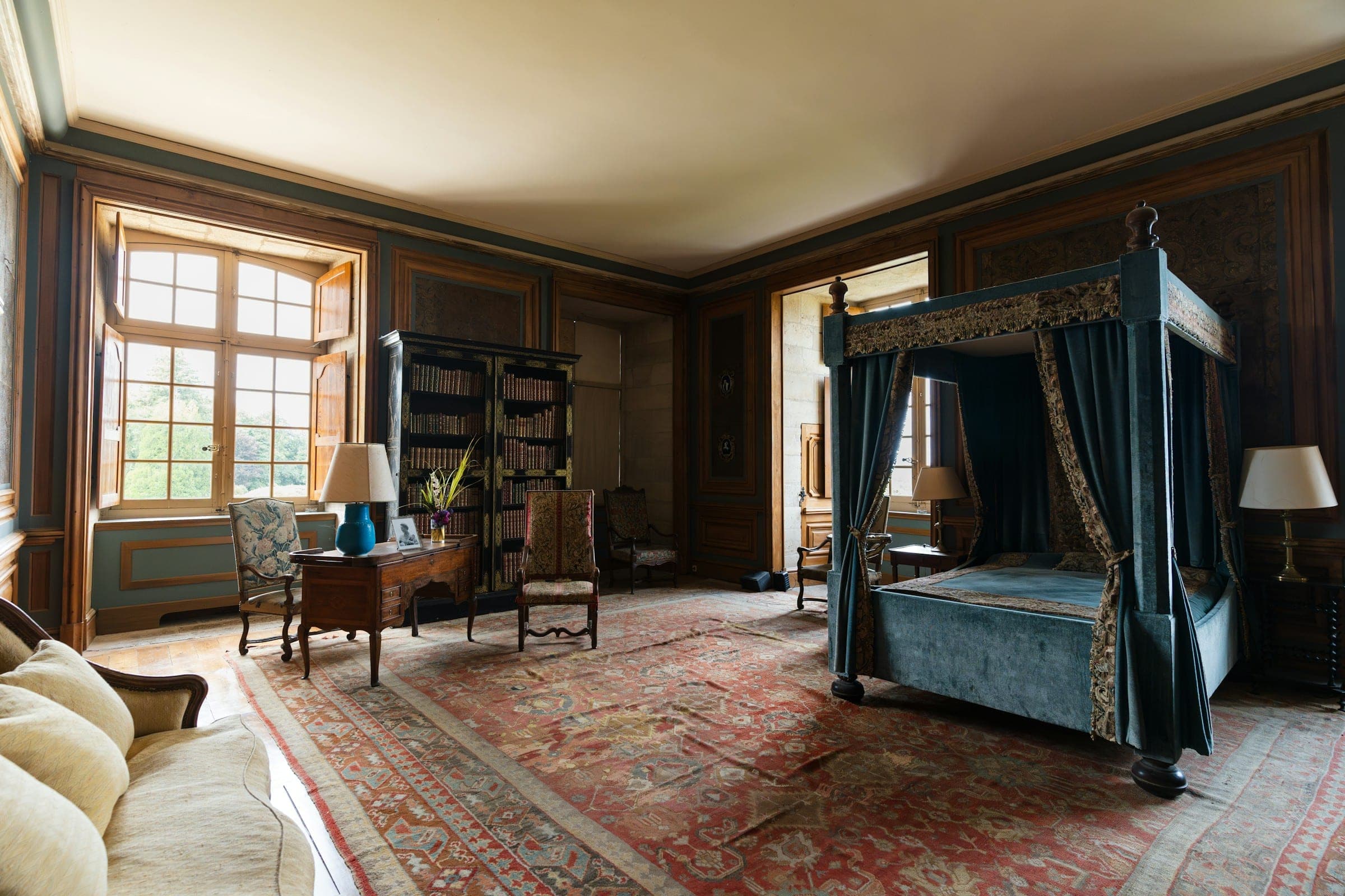 Panelled heritage bedroom with a blue canopy bed and a large faded Persian rug in soft window light